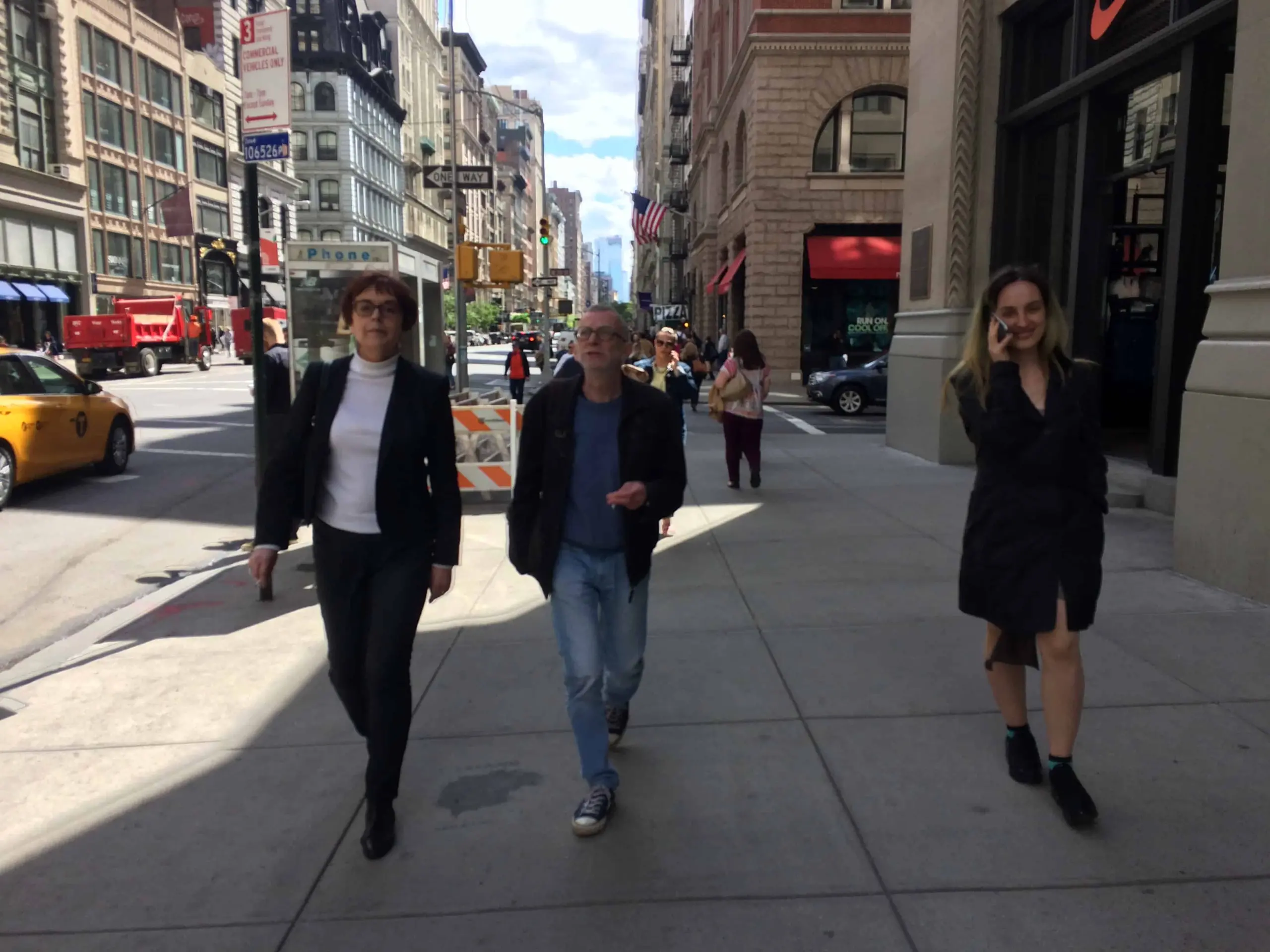Three people walk on a busy city sidewalk lined with tall buildings. A yellow taxi and pedestrians are visible in the background under a partly cloudy sky.