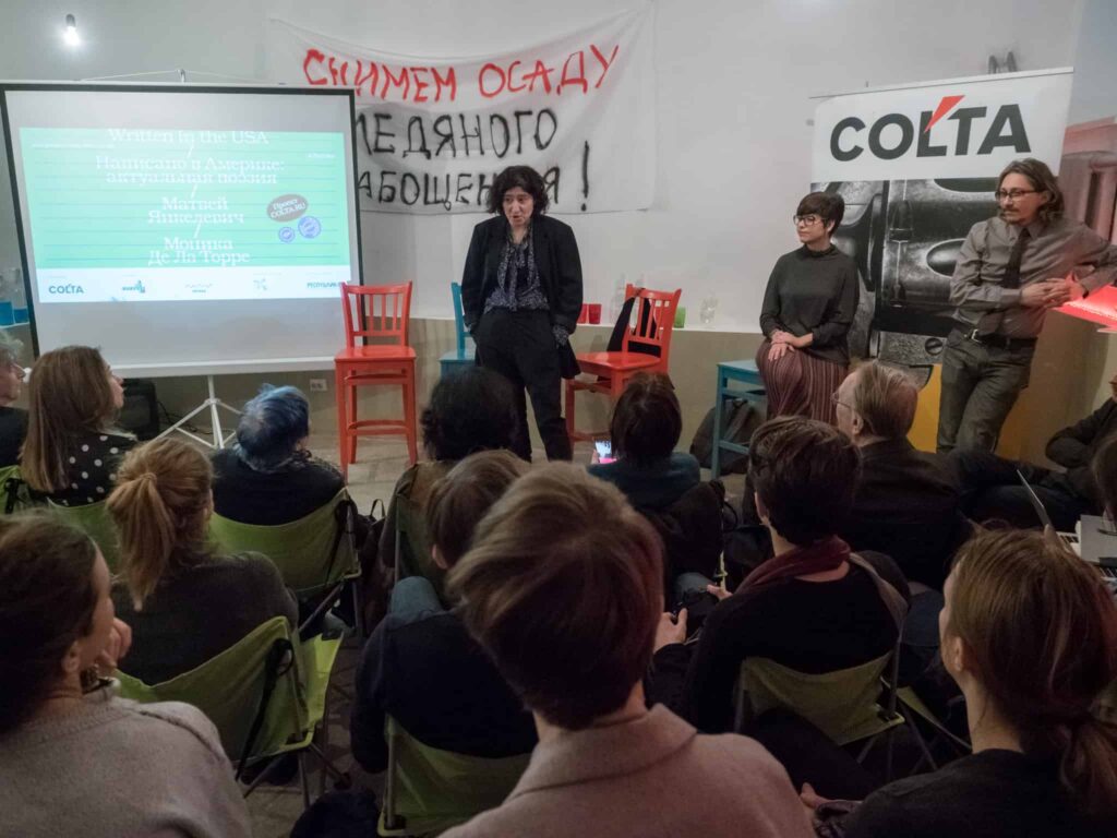 A group of people attend a panel discussion. Four speakers stand at the front near a screen displaying event information. Audience members are seated and watching. A banner with Cyrillic text hangs behind the speakers.