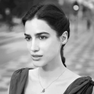 Black and white portrait of a young woman with dark hair pulled back, wearing a sleeveless top and a delicate necklace, standing outdoors on blurred, patterned pavement—capturing the quiet elegance found in Pen America Literary Awards moments.