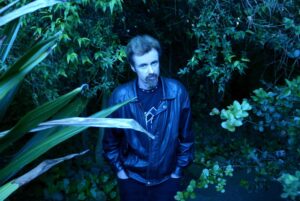A man with a beard wearing a black leather jacket stands in lush greenery, surrounded by leaves, bathed in blue-toned light, looking up at the camera with his hands in his pockets.