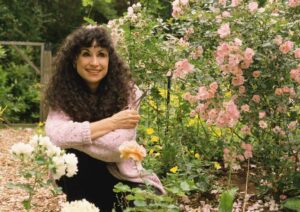 A woman with long curly hair, wearing a pink sweater, sits smiling among blooming pink and white flowers in a lush garden. Trees and greenery are visible in the background.