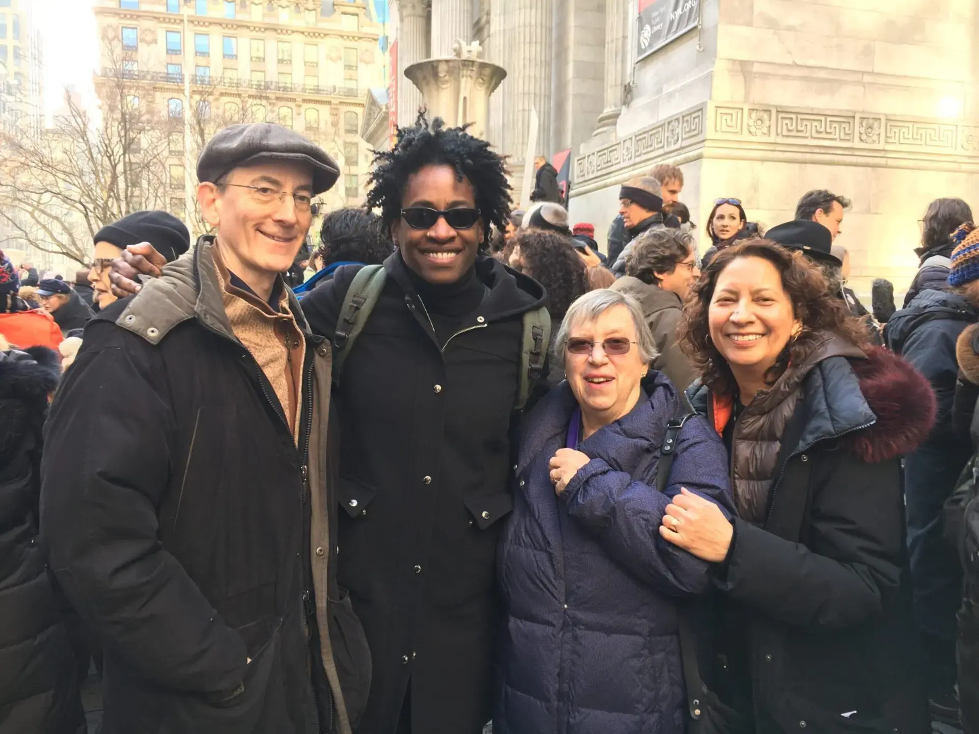 Four people stand close together, smiling at the camera in an outdoor crowd. They are wearing winter coats. Buildings and more people are visible in the background, suggesting a city setting or public event.