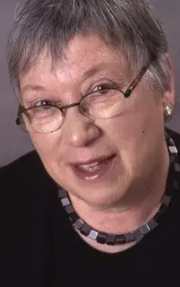 An older woman with short gray hair, wearing glasses, earrings, and a square beaded necklace, smiles at the camera against a neutral background.