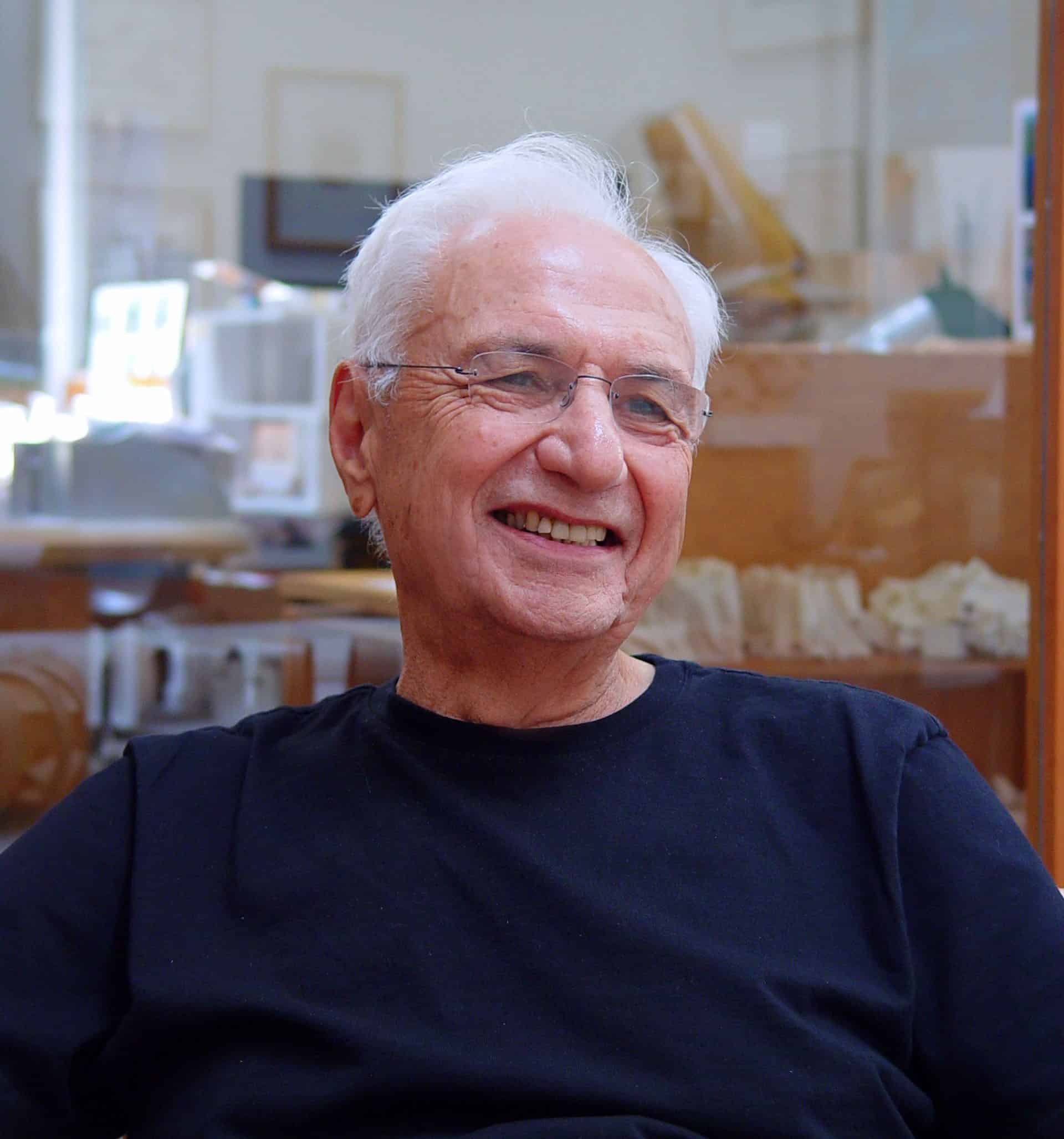 An elderly man with white hair and glasses smiles while seated indoors, wearing a black t-shirt. The background, reminiscent of Frank Gehry's creative spaces, shows shelves and blurred objects.