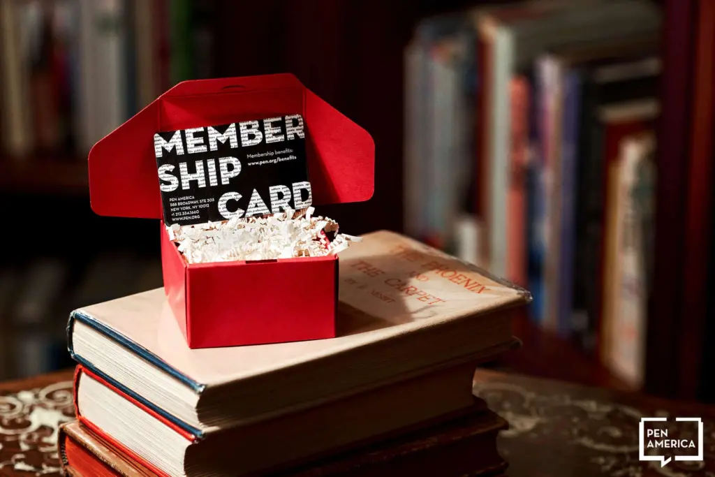 A red box containing a membership card sits open on top of stacked books, with shredded paper inside the box. A bookshelf filled with more books is blurred in the background. The Pen America logo is visible in the corner.