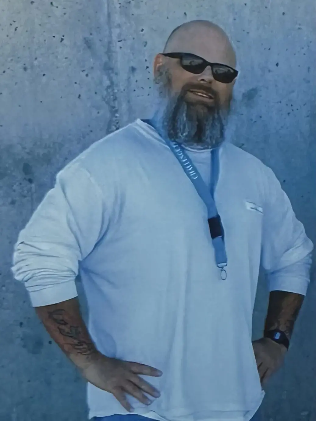A bearded man wearing sunglasses, a white long-sleeve shirt, and a blue lanyard stands with his hands on his hips in front of a gray concrete wall. He has tattoos on his forearms and is smiling slightly.