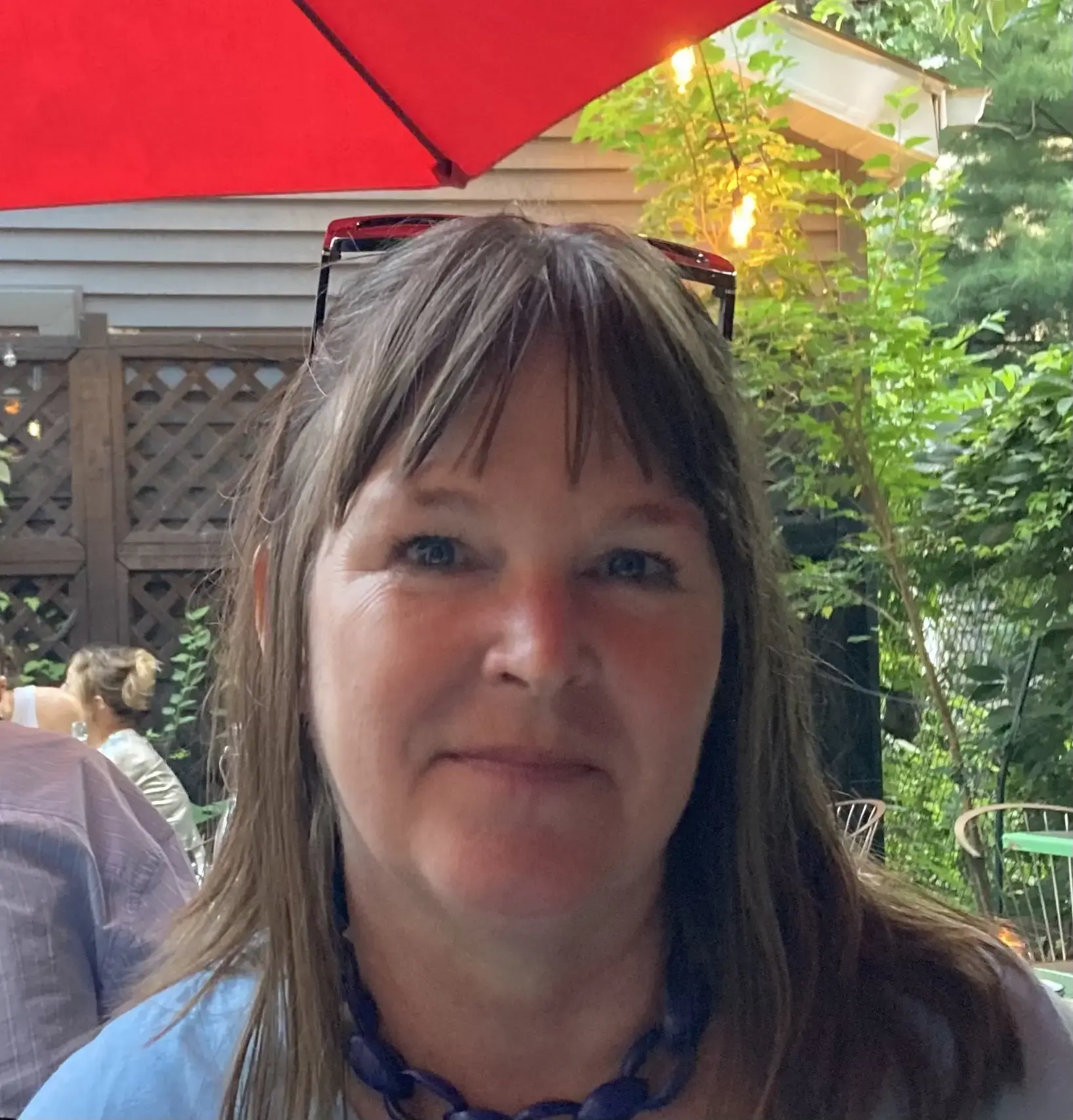 A woman with straight brown hair and bangs smiles slightly at an outdoor restaurant. She wears a blue necklace and sunglasses resting on her head. Green plants and a wooden fence are visible in the background under a red umbrella.