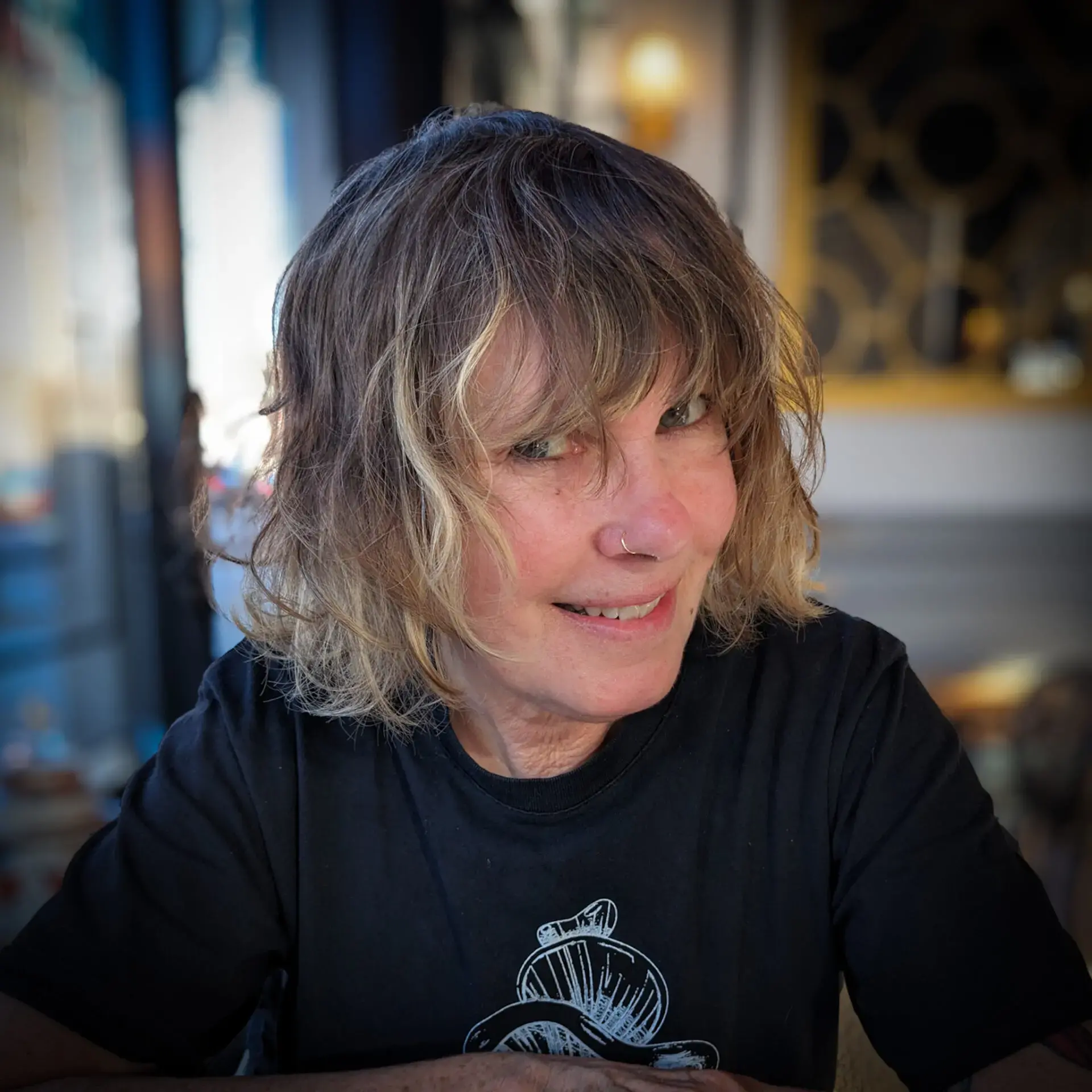 A person with wavy, chin-length hair and a nose ring smiles at the camera. They are wearing a black T-shirt and are seated indoors with blurred background decor and warm lighting.