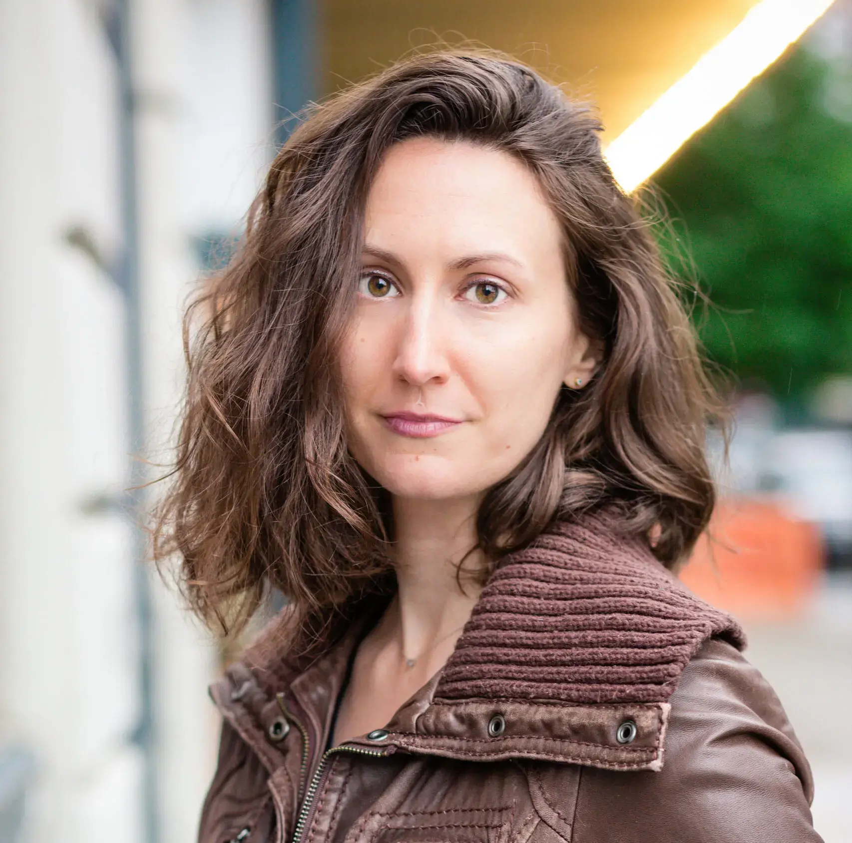 A woman with wavy brown hair and light eyes wears a brown jacket and looks at the camera with a neutral expression. The background is softly blurred, showing an outdoor urban setting.
