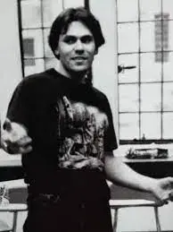A young man with dark hair stands indoors, wearing a printed t-shirt and gesturing with his hands. Behind him are large windows with a grid pattern. The image is black and white.