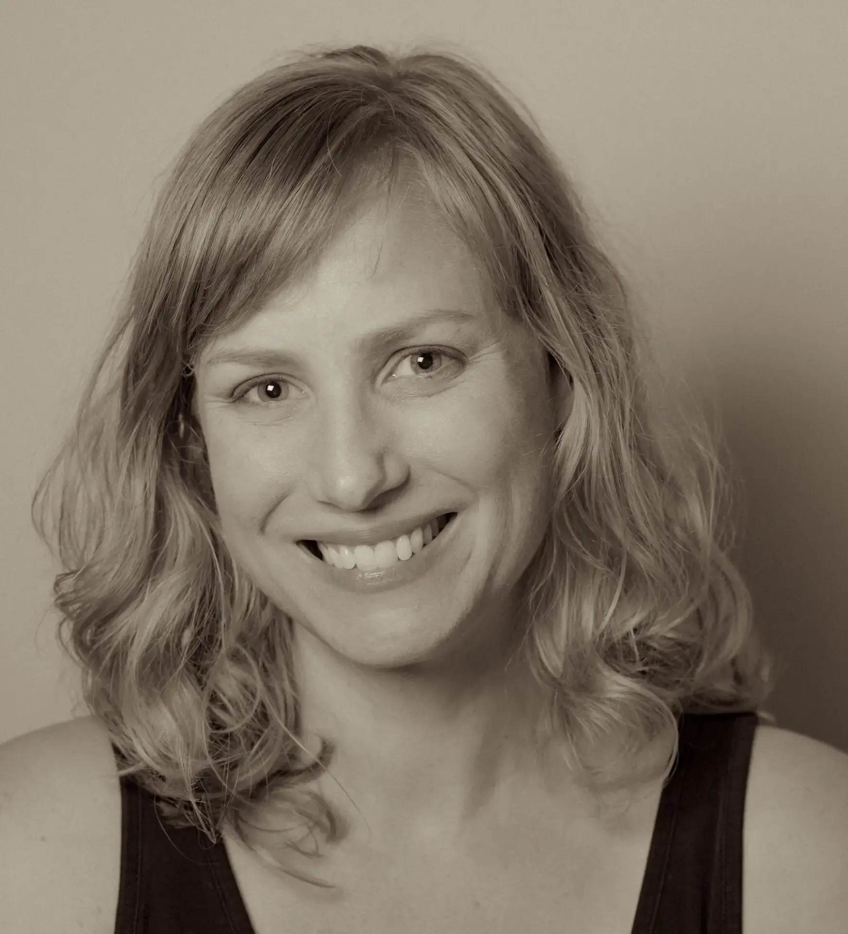 A woman with wavy, shoulder-length blonde hair smiles at the camera. She is wearing a sleeveless dark top, and the photo is in sepia tone.