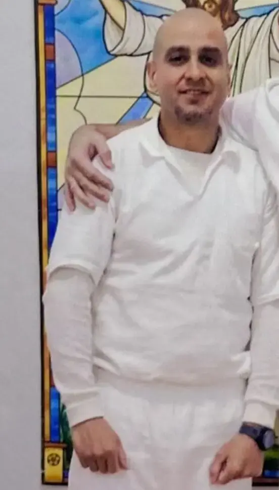 A man with a shaved head, dressed in all white, stands in front of a colorful stained glass-style background featuring religious imagery. Another person’s arm is resting on his shoulder, but that person is mostly out of frame.