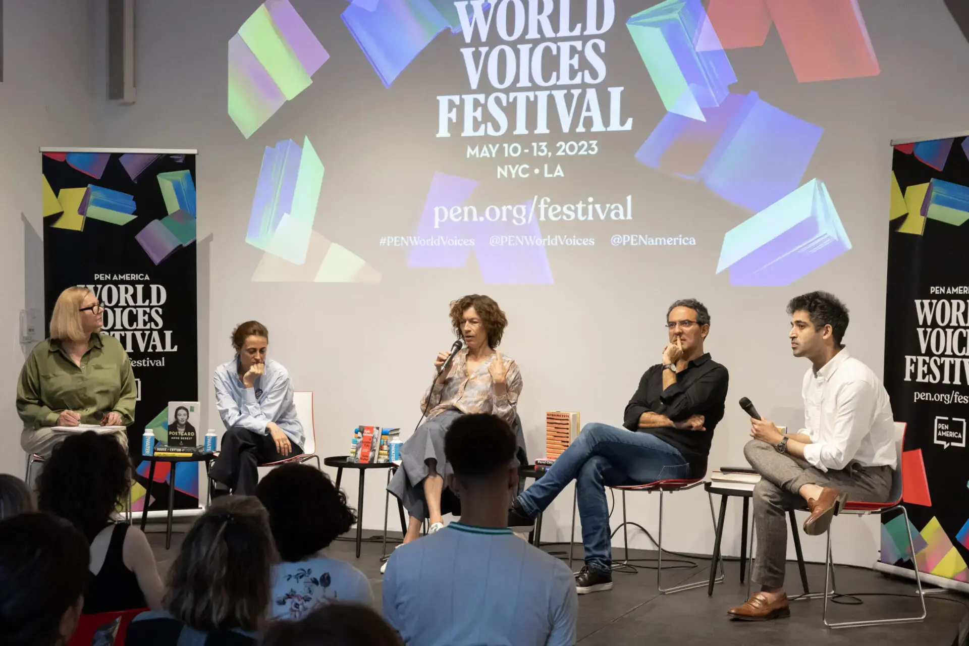 Five panelists sit on stage having a discussion at the PEN America World Voices Festival 2023, as the audience watches. A large screen behind them displays the event name and details.
