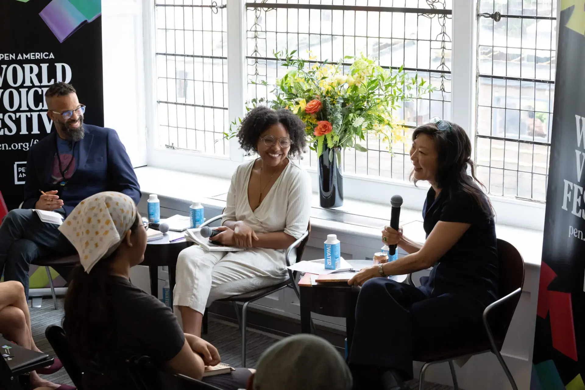 Three people sit on chairs in front of an audience, engaging in a World Voices Festival 2023 panel discussion. One woman holds a microphone while speaking. Large windows and a vase of flowers are seen in the background.