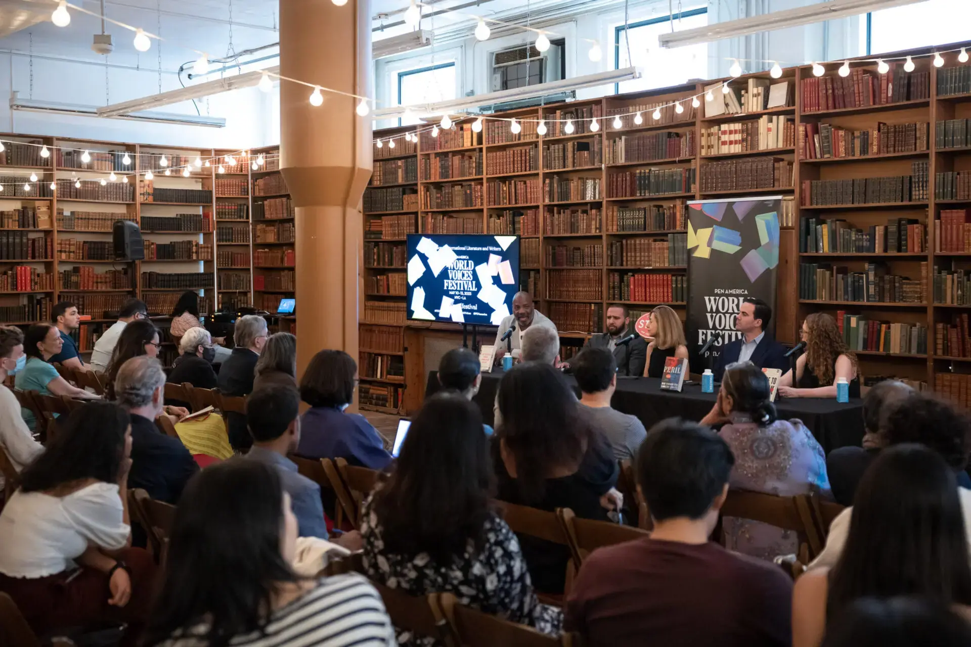 A panel of speakers sits at a table in front of an audience in a library, surrounded by bookshelves and string lights, with a screen displaying World Voices Festival 2023.