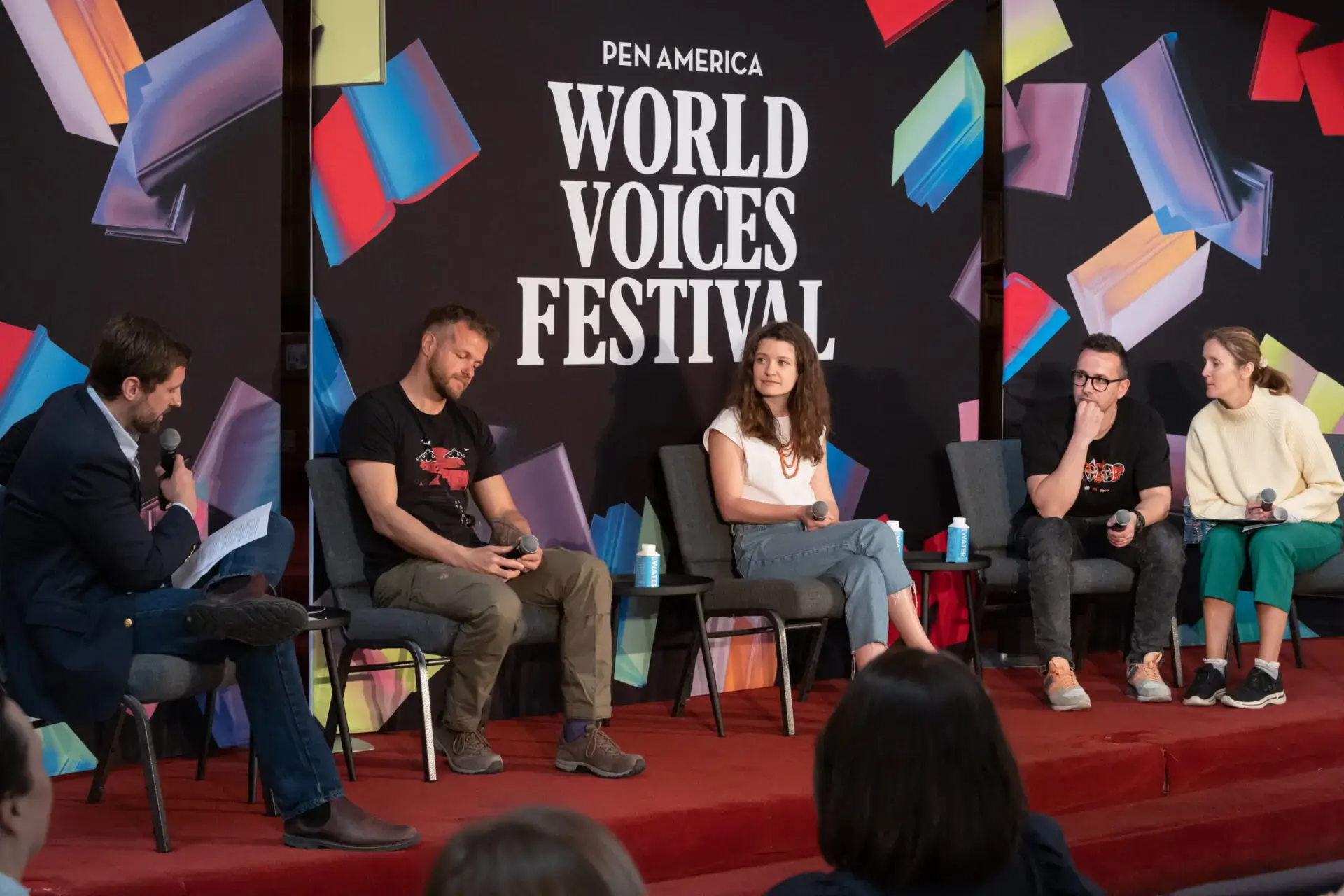 Five people sit onstage in front of a World Voices Festival 2023 backdrop, engaged in a lively panel discussion. Four are seated in a row while one on the left holds a microphone and paper. Audience members’ heads are visible in the foreground.