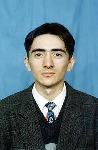 A young man with short dark hair, wearing a dark checked suit, patterned tie, and gray sweater, poses against a plain blue background.