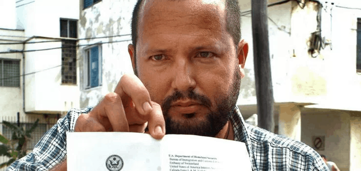 A man with a beard and plaid shirt holds up an official document displaying a U.S. government seal, standing outdoors in front of weathered buildings and a fence.