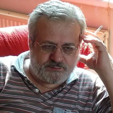 A middle-aged man with gray hair and a beard, wearing glasses and a striped polo shirt, sits on a red chair, holding a cigarette and looking thoughtful or concerned. Sunlight shines on his face.