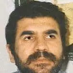 A man with dark hair and a thick beard looks upwards. He is positioned in front of a light background with some indistinct shapes or objects behind him.