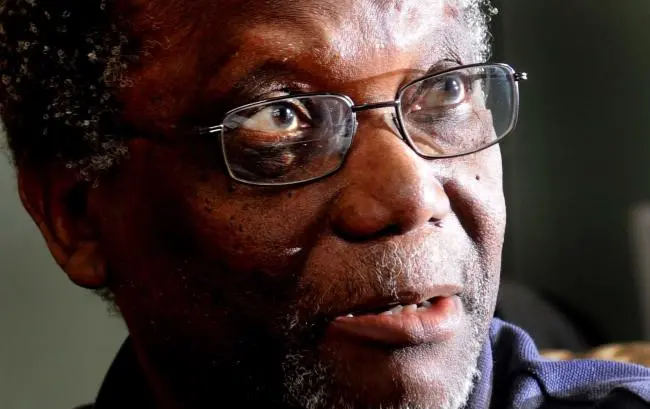 Close-up of an older man with glasses, gray hair, and a short beard, looking slightly upwards with a thoughtful expression. Sunlight is illuminating part of his face.