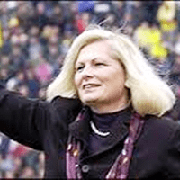 A woman with blonde hair wearing a dark coat and scarf stands outdoors, smiling and raising her arm, with a blurred crowd in the background.