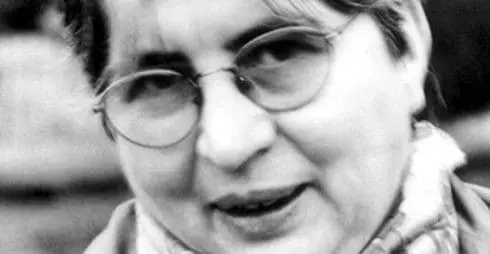 Black and white close-up photo of an older person with short hair and glasses, wearing a scarf and looking slightly past the camera.