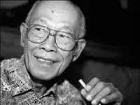 An elderly man with glasses smiles while holding a cigarette. He is wearing a patterned shirt and appears to be looking slightly to the side. The image is in black and white.