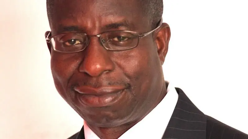 A man wearing glasses and a dark pinstripe suit with a white shirt, smiling slightly, photographed against a plain white background.