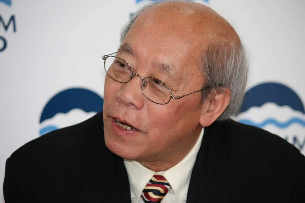 An older man with glasses and thinning hair, wearing a black suit, white shirt, and a patterned tie, speaks while looking slightly to the side. A blurred logo is visible in the background.