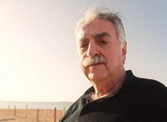 An older man with gray hair and a mustache stands outdoors near a beach, wearing a dark shirt. The sky is clear and bright, with the ocean visible in the background.