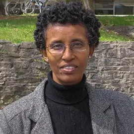A woman with short curly hair and glasses, wearing a gray coat and black turtleneck, stands outdoors in front of a stone wall and grassy area with bicycles in the background.