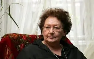 An older woman with short, curly brown hair and glasses sits on a patterned chair in front of sheer white curtains, wearing a dark top and looking slightly to the side.