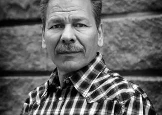 A middle-aged man with a mustache wearing a plaid shirt stands in front of a textured stone wall, looking directly at the camera in this black and white photo.