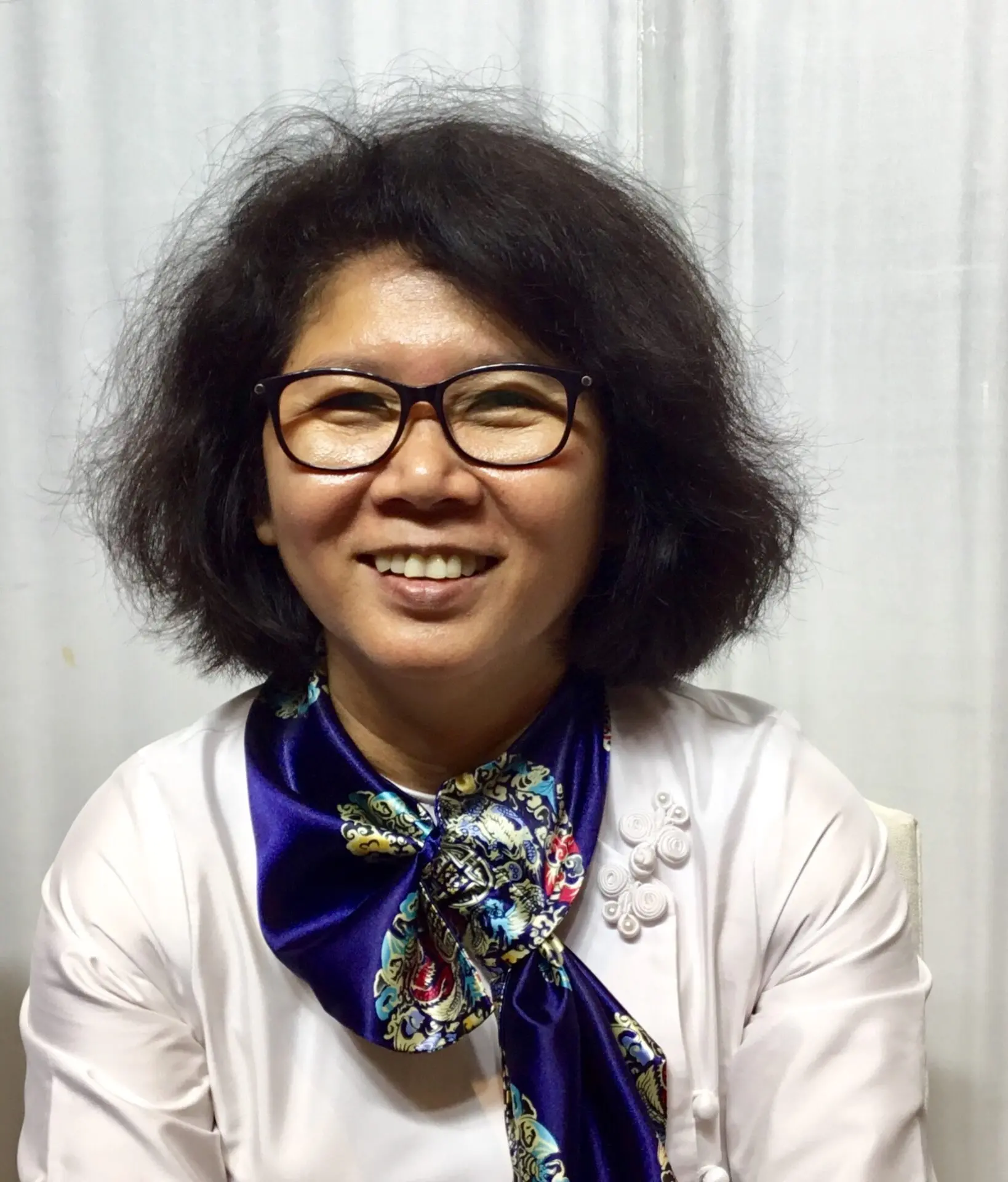 A woman with short, dark hair and glasses smiles at the camera. She is wearing a white blouse with decorative buttons and a blue floral scarf. The background is plain and light-colored.