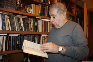 An elderly man wearing a gray sweater stands in front of bookshelves, reading an open book. The shelves behind him are filled with various books.