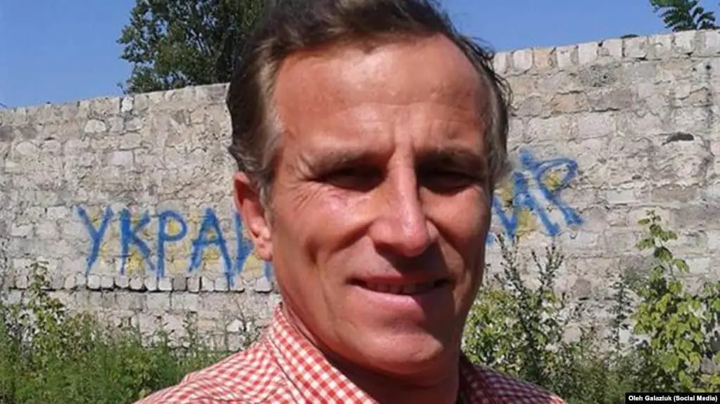 A man in a red checkered shirt taking a selfie outdoors, standing in front of a stone wall with blue graffiti text in Cyrillic script and greenery around him.