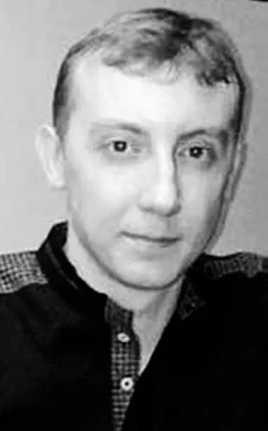 A black and white photo of a man with short, light-colored hair, wearing a dark shirt with a patterned collar, looking at the camera with a neutral expression.