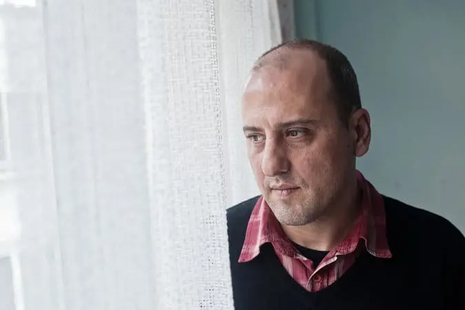 A man with a shaved head wearing a red plaid shirt and dark sweater gazes thoughtfully out of a window, partially covered by a sheer white curtain, in a softly lit room.