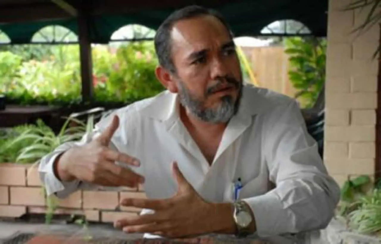 A man with a beard, wearing a white shirt and a watch, sits at a table outdoors with plants in the background, gesturing with his hands while talking.