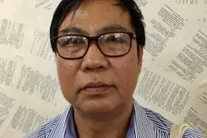 A man wearing glasses and a blue-striped shirt looks at the camera. Pages with printed text are displayed on the wall behind him.