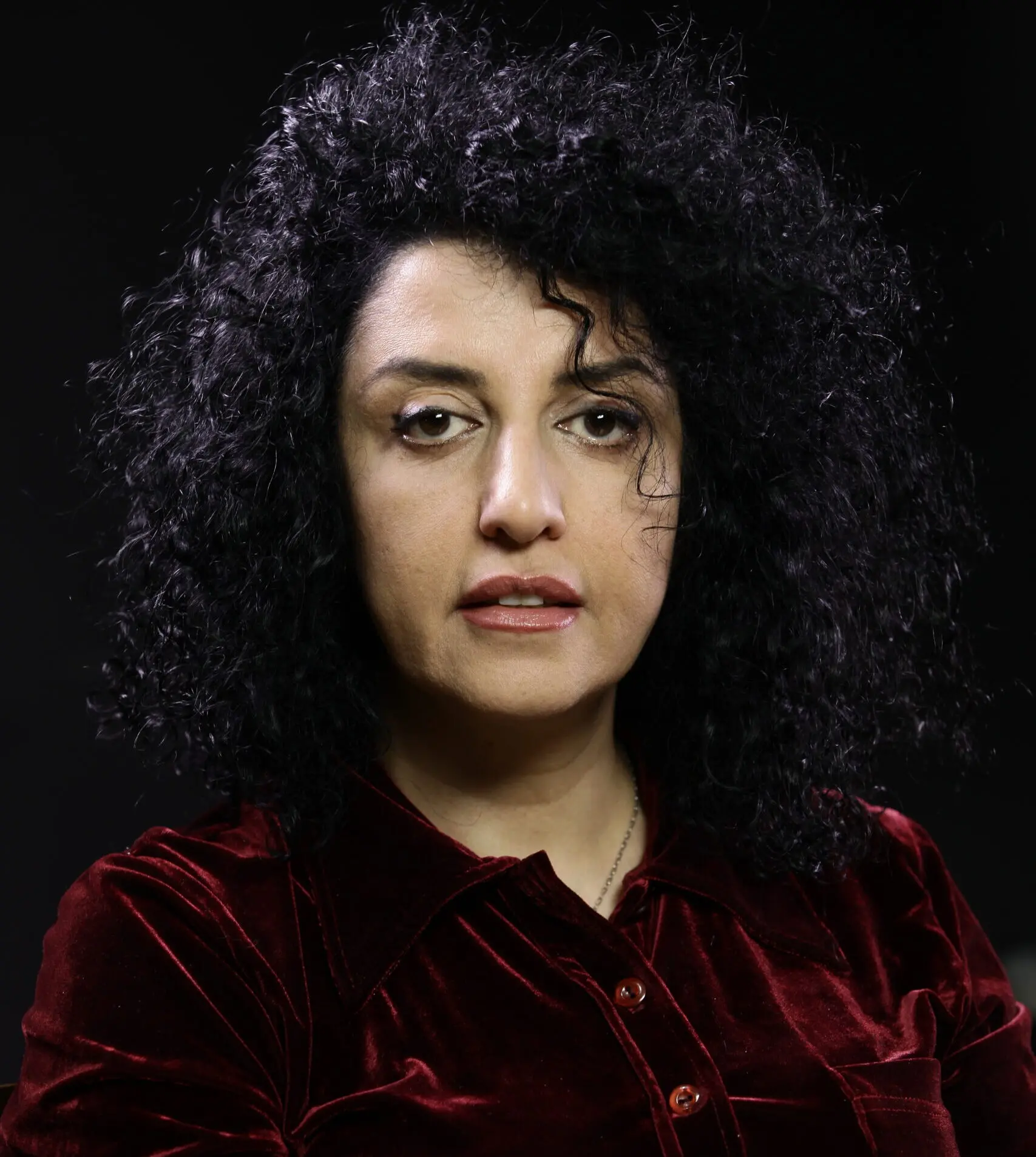 A woman with curly black hair wearing a dark red velvet shirt sits against a dark background, looking directly at the camera with a neutral expression, reminiscent of portraits of Narges Mohammadi.