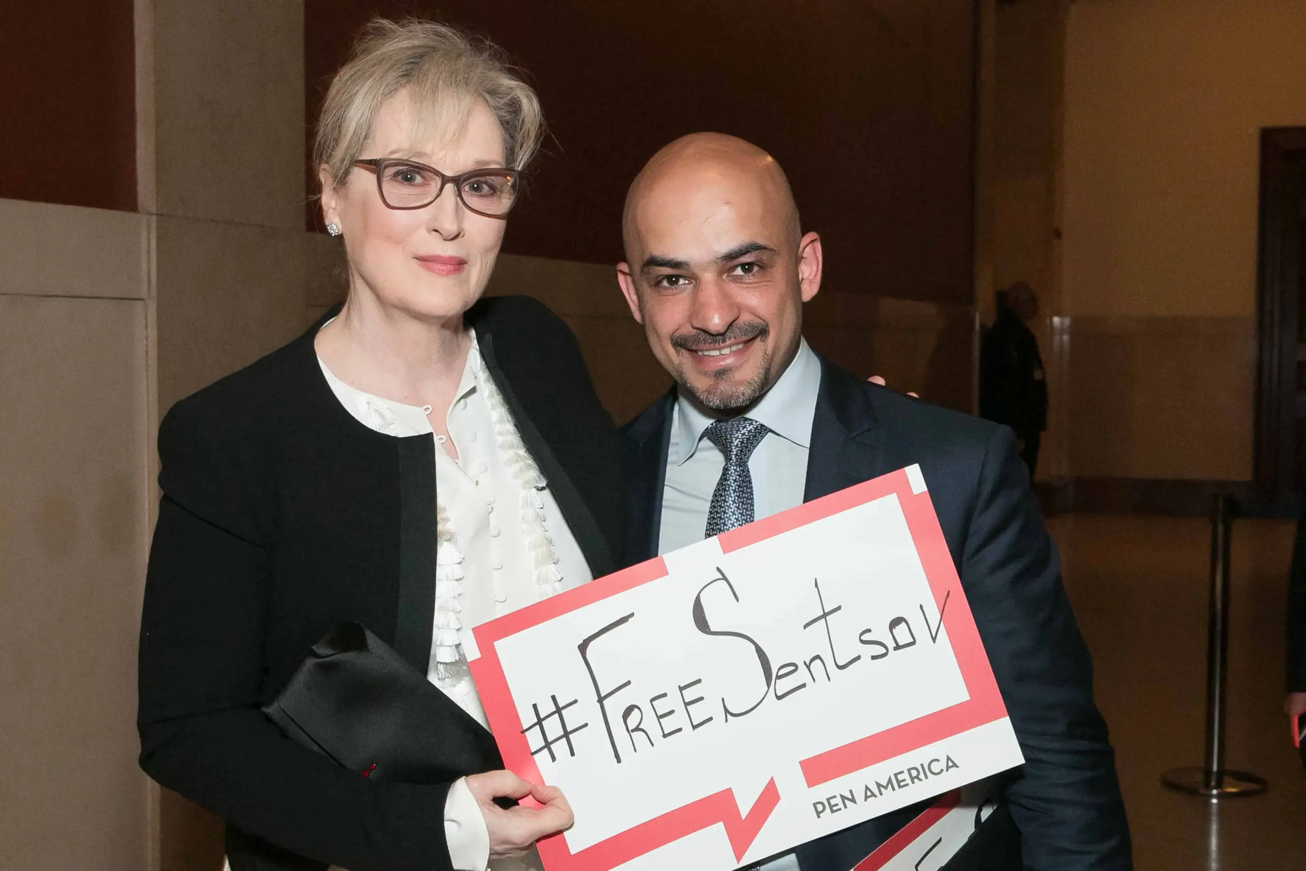 A woman and a man stand together smiling indoors, dressed formally. The woman holds a sign that reads #FreeSentsov PEN America, showing support for filmmaker Oleg Sentsov.