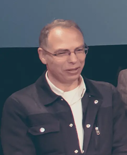 A middle-aged man with short hair and glasses is wearing a dark jacket over a white shirt, standing in front of a plain blue and black background.