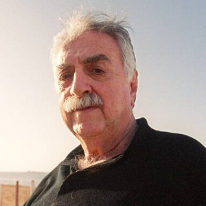 An older man with gray hair and a mustache stands outdoors, wearing a dark shirt. The sky is clear and bright behind him, suggesting it is daytime.