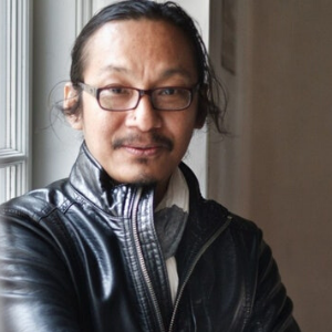 A man with long dark hair tied back, wearing glasses and a black leather jacket, stands indoors near a window, looking at the camera with a slight smile.