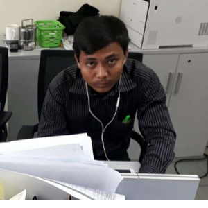 A man in a black striped shirt sits at a desk with papers, wearing earphones and looking up at the camera. Office furniture, a printer, and a green basket are in the background.