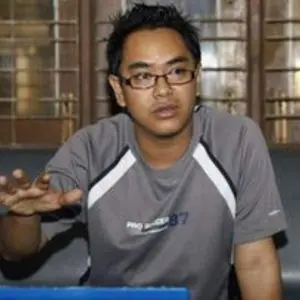 A man wearing glasses and a grey t-shirt sits on a couch, gesturing with his right hand as he speaks. There are wooden window panels in the background.