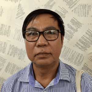 A man wearing glasses and a blue striped shirt stands in front of a wall covered with printed pages arranged in a collage. He is looking directly at the camera with a neutral expression.