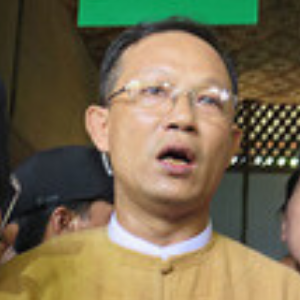 A man wearing glasses and a tan traditional outfit stands indoors with his mouth slightly open, surrounded by other people in the background.